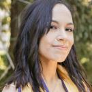 Headshot of young person with straight black hair just past shoulders, grinning and wearing gold graduation stole, green trees in sun in background